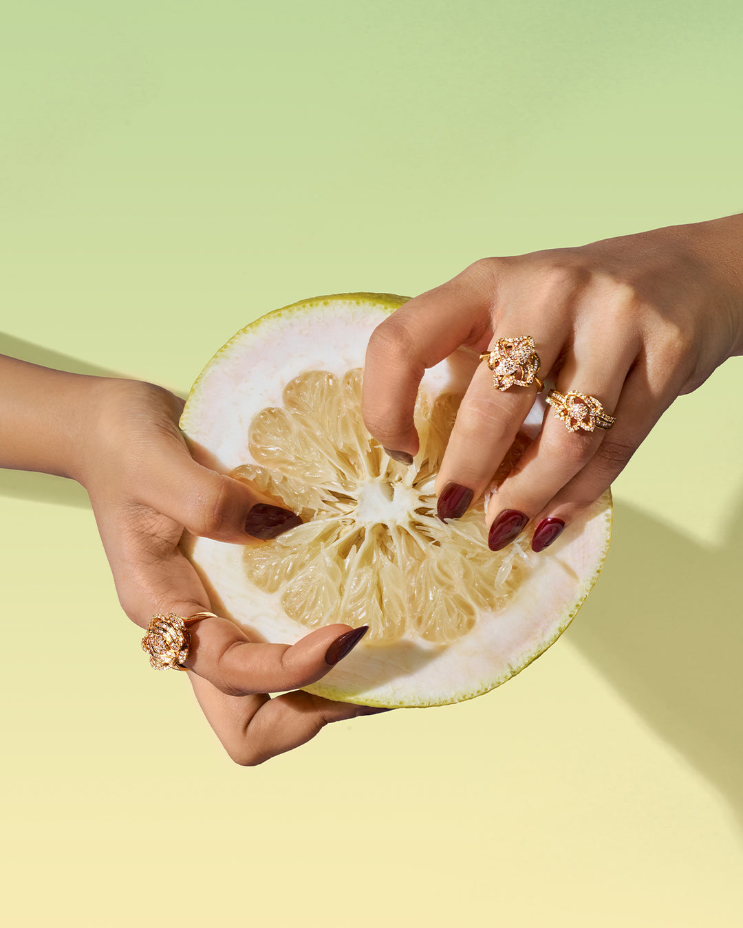 Two hands wearing rings, holding a halved grapefruit against a pale green backdrop