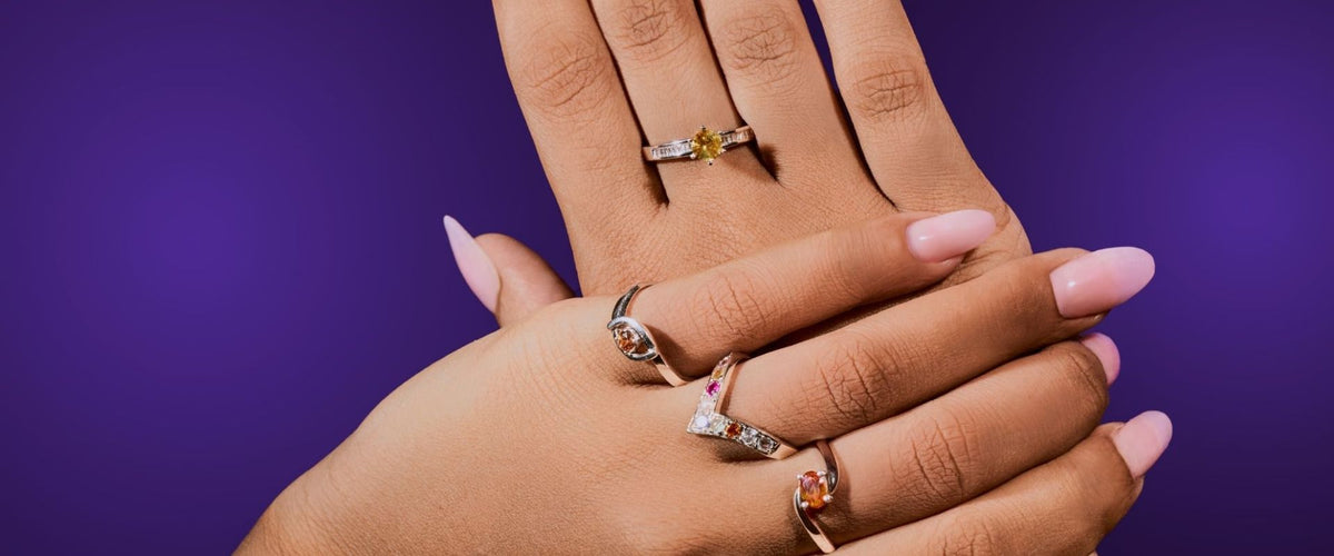Two hands resting together, wearing gemstone rings, on a violet backdrop